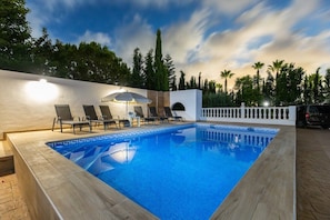 En rektangulär pool med ett geometriskt mönster av kakel, omgiven av solstolar och ett parasoll, med ett vitt staket och träd i bakgrunden.