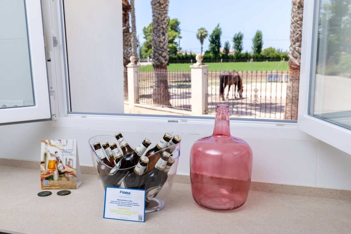 A window with a view of a fenced area, a bottle of pink liquid, and a container of beer bottles.