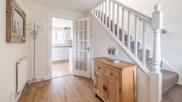 Een hal met houten vloer, een witte radiator, een witte deur, een houten kast en een trap met witte leuningen.