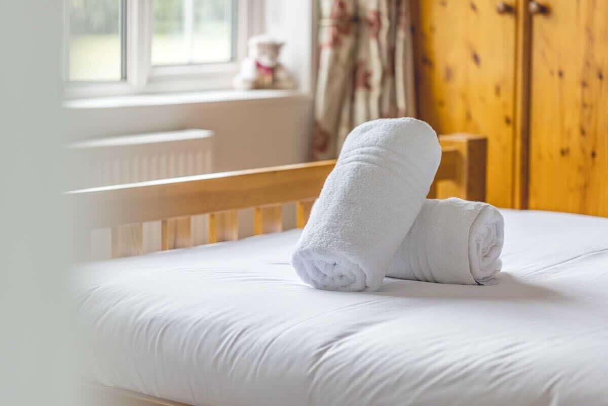 Ein ordentlich bezogenes Bett mit weißen Leinen und einem aufgerollten weißen Handtuch am Kopfteil.