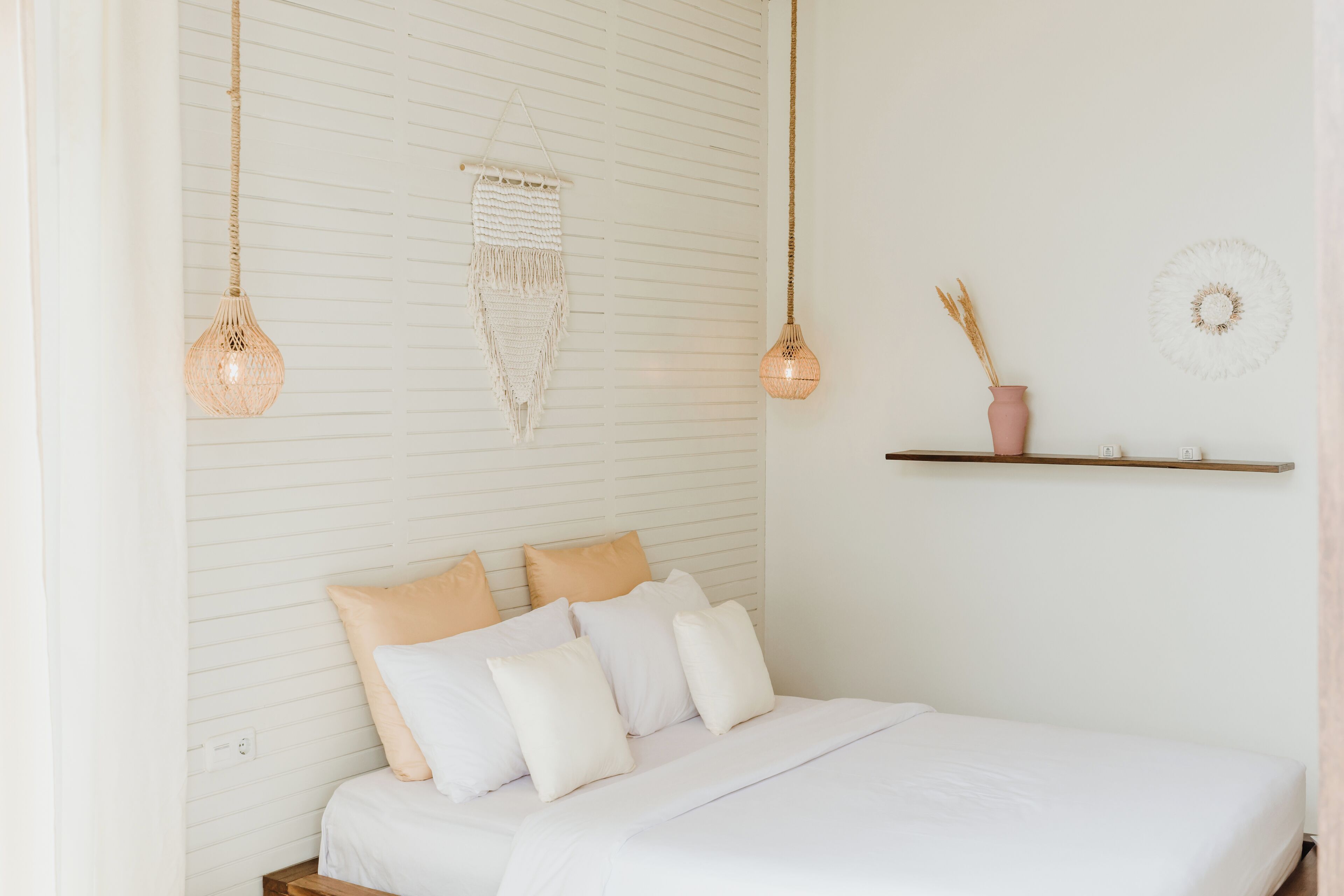 Una cama bien hecha con ropa de cama blanca y beige, un colgador de macramé en la pared y un florero rosa con plantas secas sobre una estantería.