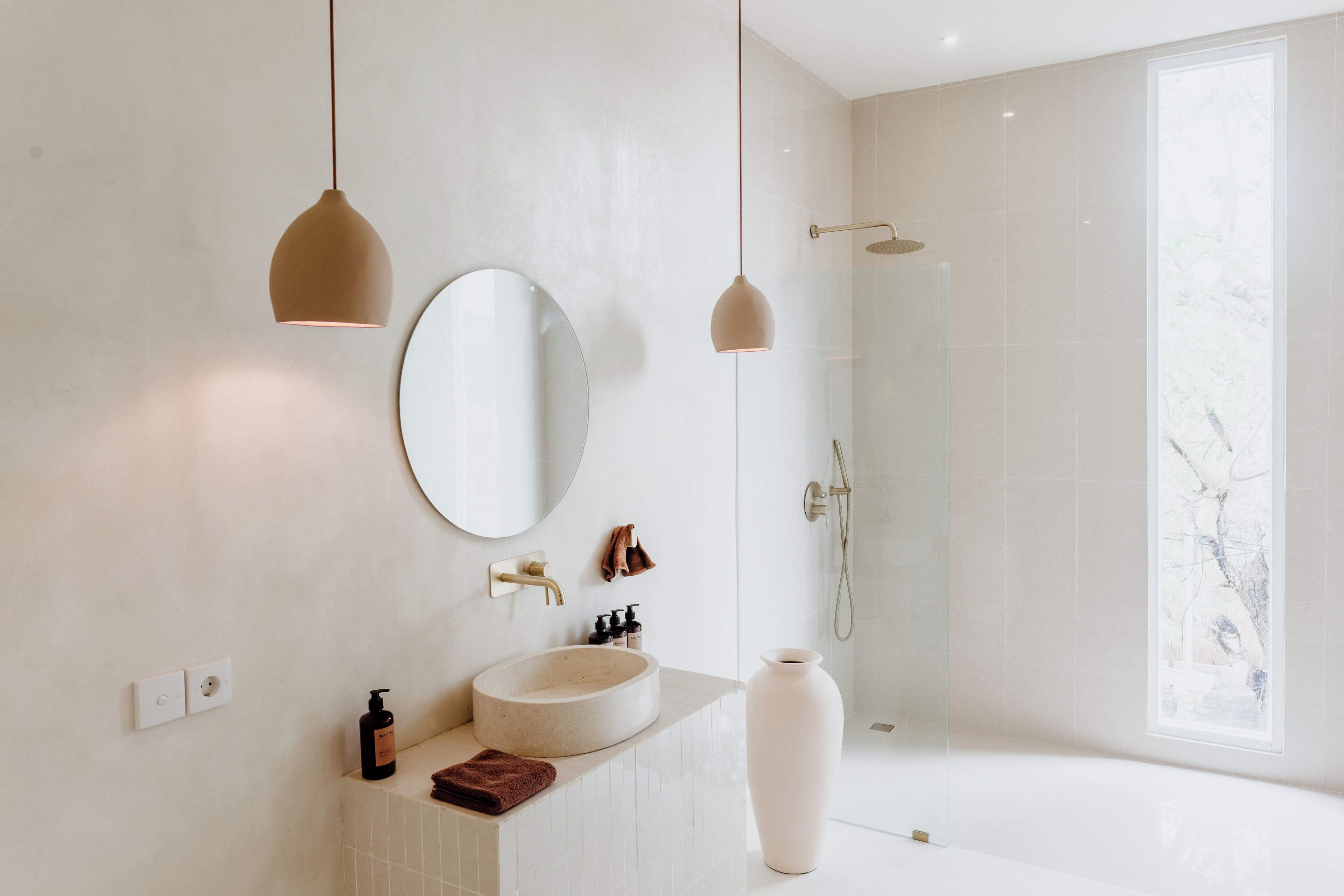 A modern bathroom with a round mirror, a sink, a large vase, and a shower.