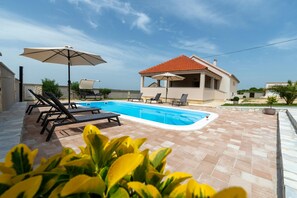 A pool area with lounge chairs and umbrellas, a house with a red roof, and a view of the sea. - Villa Zrilić ZadarVillas (Perušić Benkovački)