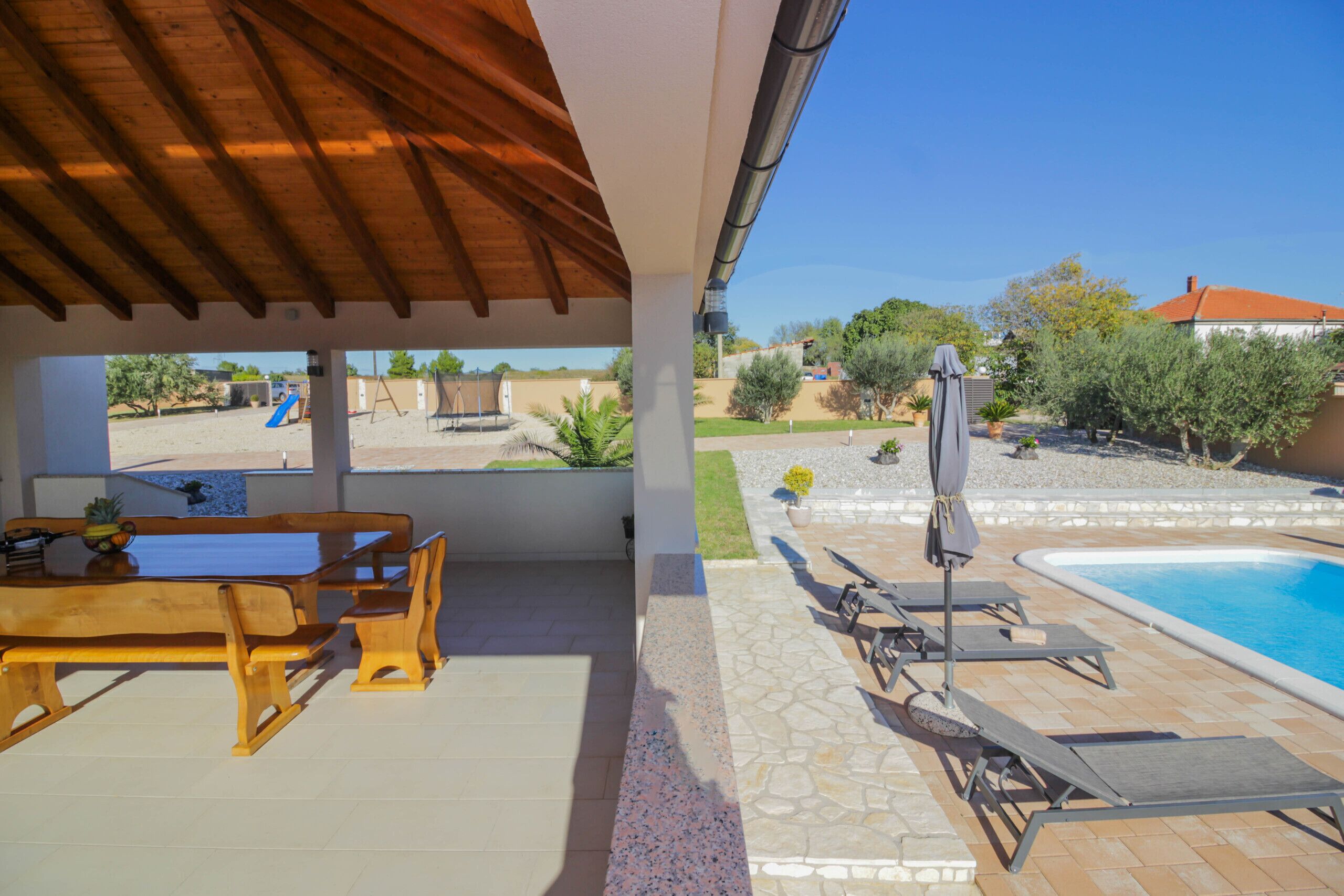 Portico coperto con tavolo e sedie in legno, zona piscina con lettini e vista sul giardino.
