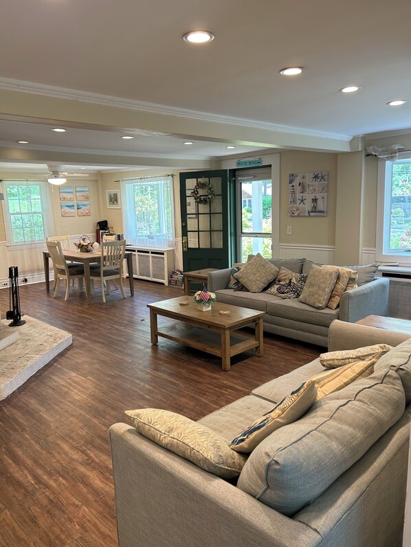 A spacious living room with a wooden floor, a grey sofa, a coffee table, and a dining area with chairs and a table.