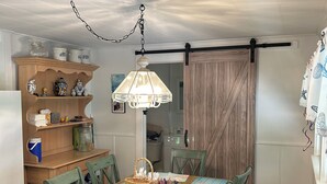 Un coin repas avec une table en bois, des chaises vertes, une lampe suspendue et une étagère en bois où sont rangés les ustensiles de cuisine.
