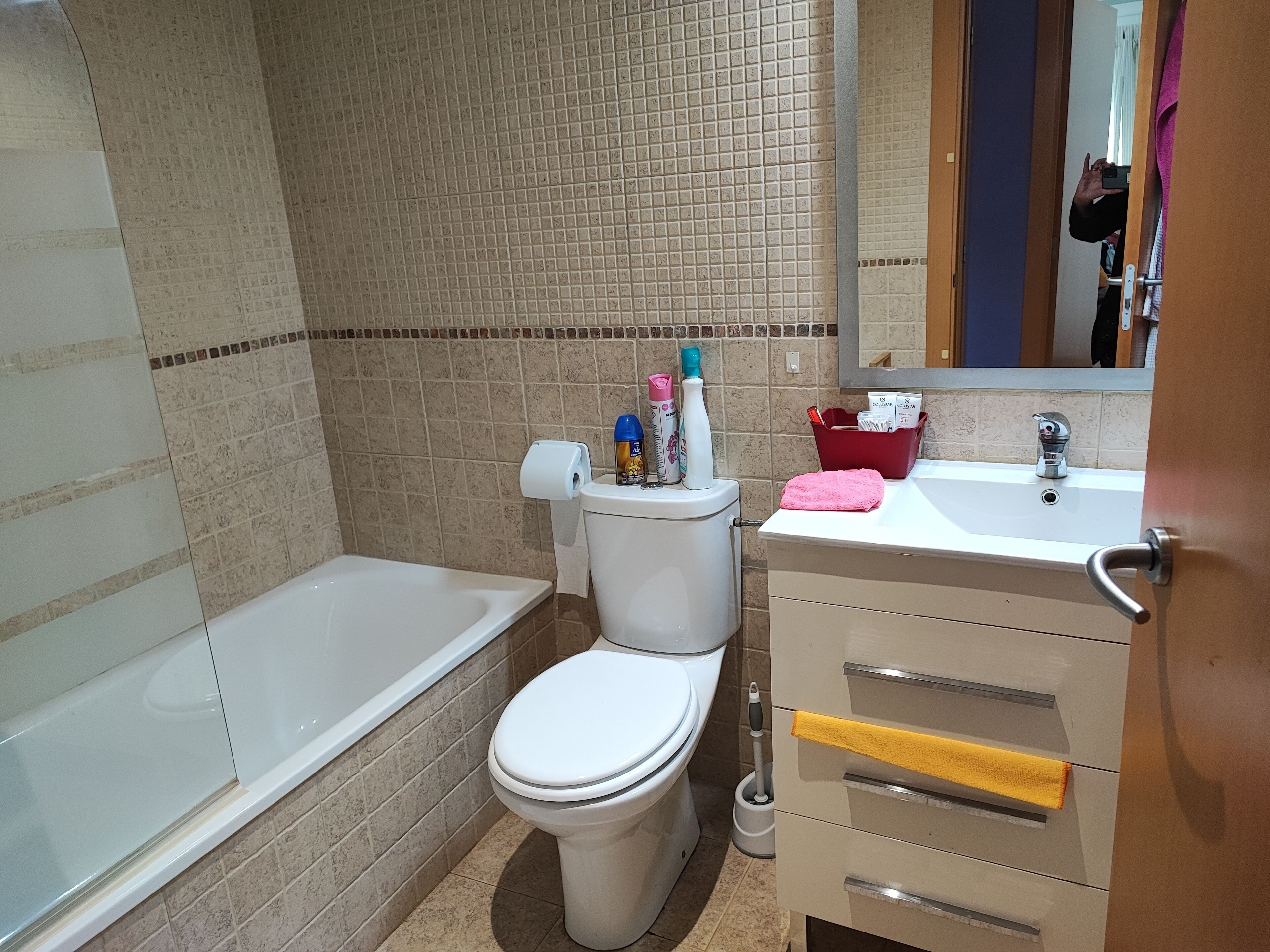 Une salle de bain avec des toilettes, un lavabo et une baignoire. On trouve des articles de toilette sur le réservoir des toilettes et un panier rouge sur le lavabo.