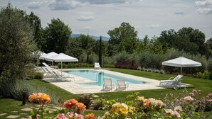 A rectangular swimming pool with white lounge chairs and umbrellas, surrounded by a well-maintained lawn and flowering plants. - Villa in Foiano Della Chiana with 6 bedrooms sleeps 14 (Foiano Della Chiana)