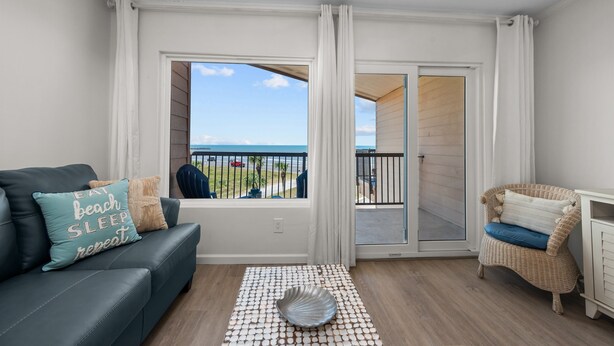 A living room with a dark sofa, a wicker chair, and a view of the beach through a sliding glass door. - New Heated Pool & Hot tub! Fantastic Ocean Views! (Galveston)