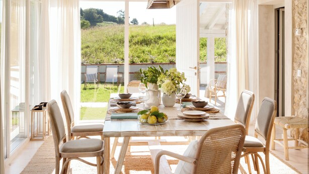 A dining area with a table set for six, surrounded by wicker chairs, with a view of a green landscape through large windows. - Casa con Jardín y Piscina Climatizada Cerca de Comillas a 10 min de la Playa (valdaliga)