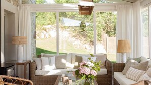 A cozy living room with wicker furniture, a coffee table with books and a vase of flowers, and large windows offering a view of the outdoors. - Casa con Jardín y Piscina Climatizada Cerca de Comillas a 10 min de la Playa (valdaliga)