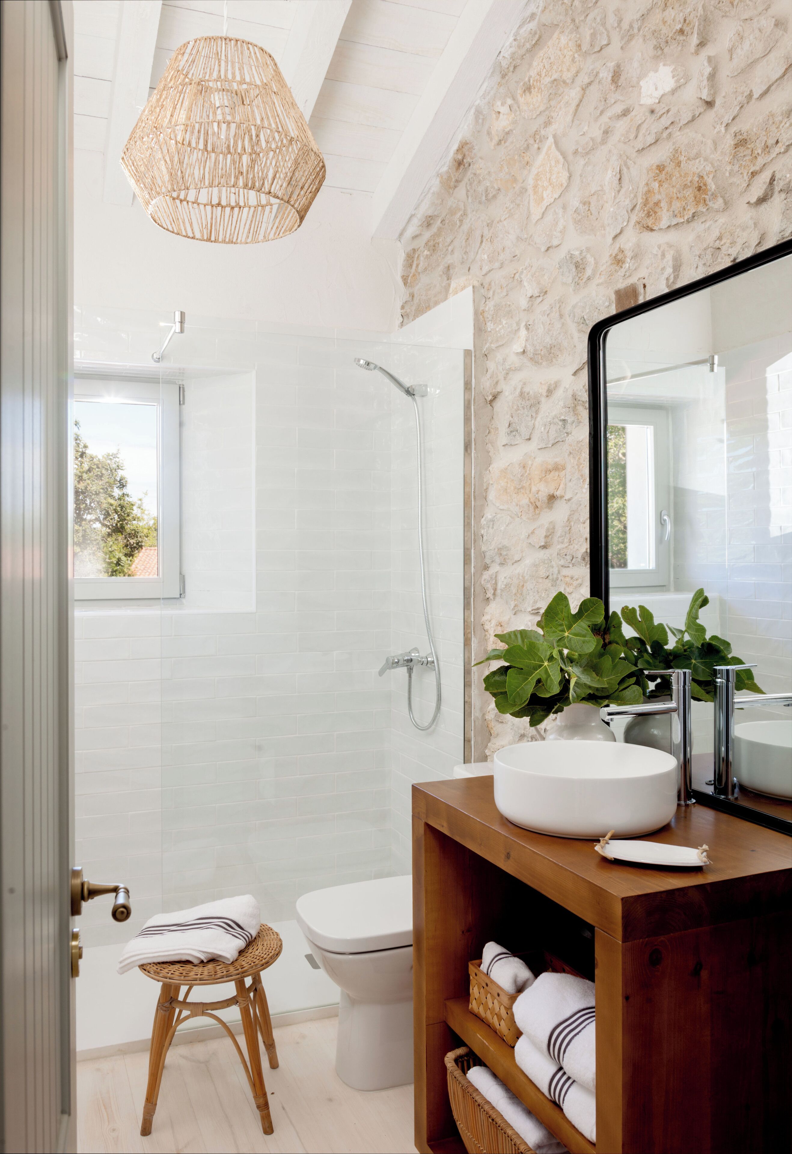 Un baño moderno con pared de piedra, un espejo grande, un lavabo blanco y un taburete de mimbre.