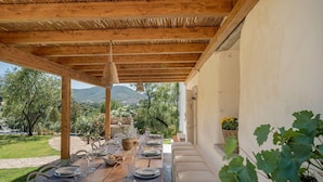 An outdoor dining area with wooden tables and chairs, a woven ceiling, and a view of a garden and mountains. - Villa Canto Antico -SARDINIA UNLIMITED (Porto San Paolo)
