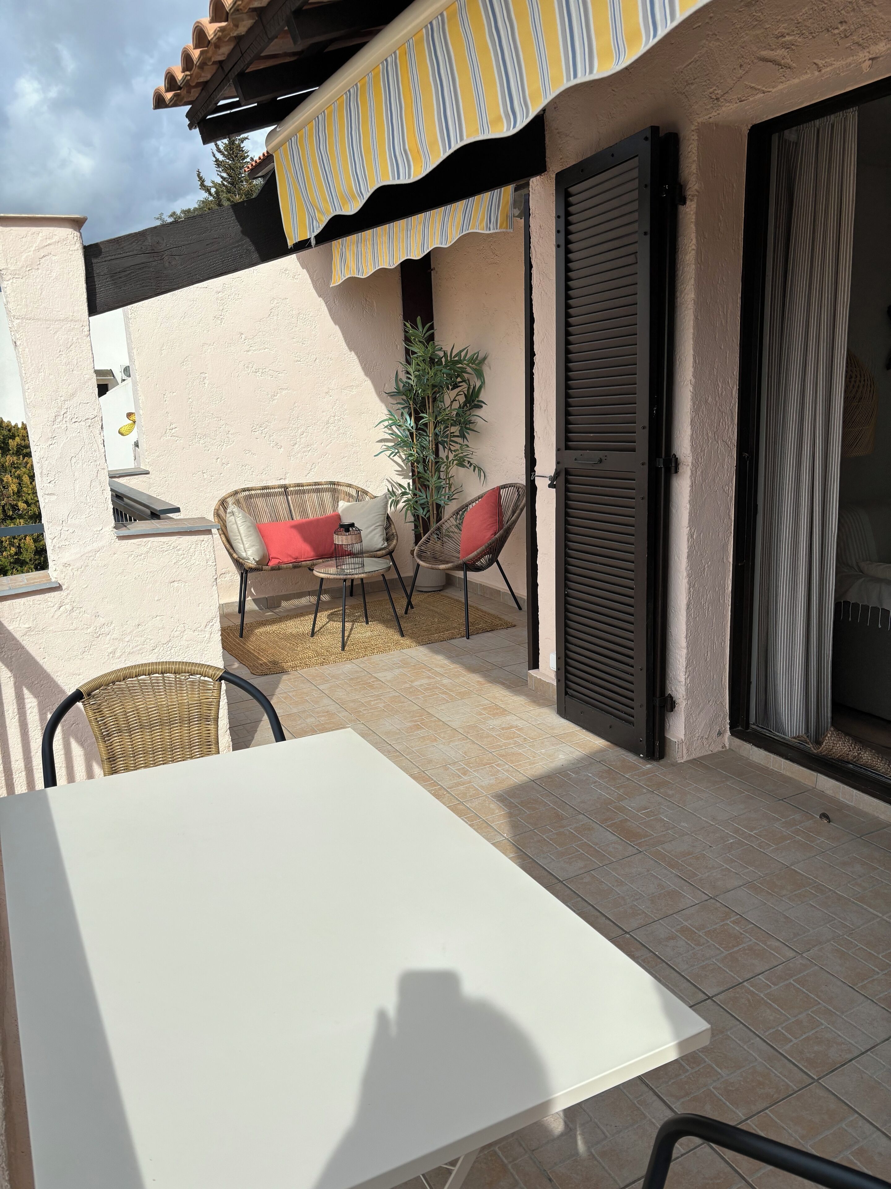 A patio with a white table and wicker chairs, a potted plant, and a striped awning.
