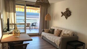 A living room with a sofa, a table, and chairs. There is a balcony with a view of buildings and a striped awning. - Magnifique VUE MER Golfe DE ST Tropez (La Croix-Valmer)