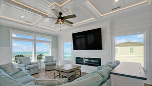 Un vaste salon comprenant un grand canapé, deux fauteuils, une table basse et un téléviseur à écran plat fixé au mur. De grandes fenêtres offrent une vue sur l’océan, et un balcon est également disponible.