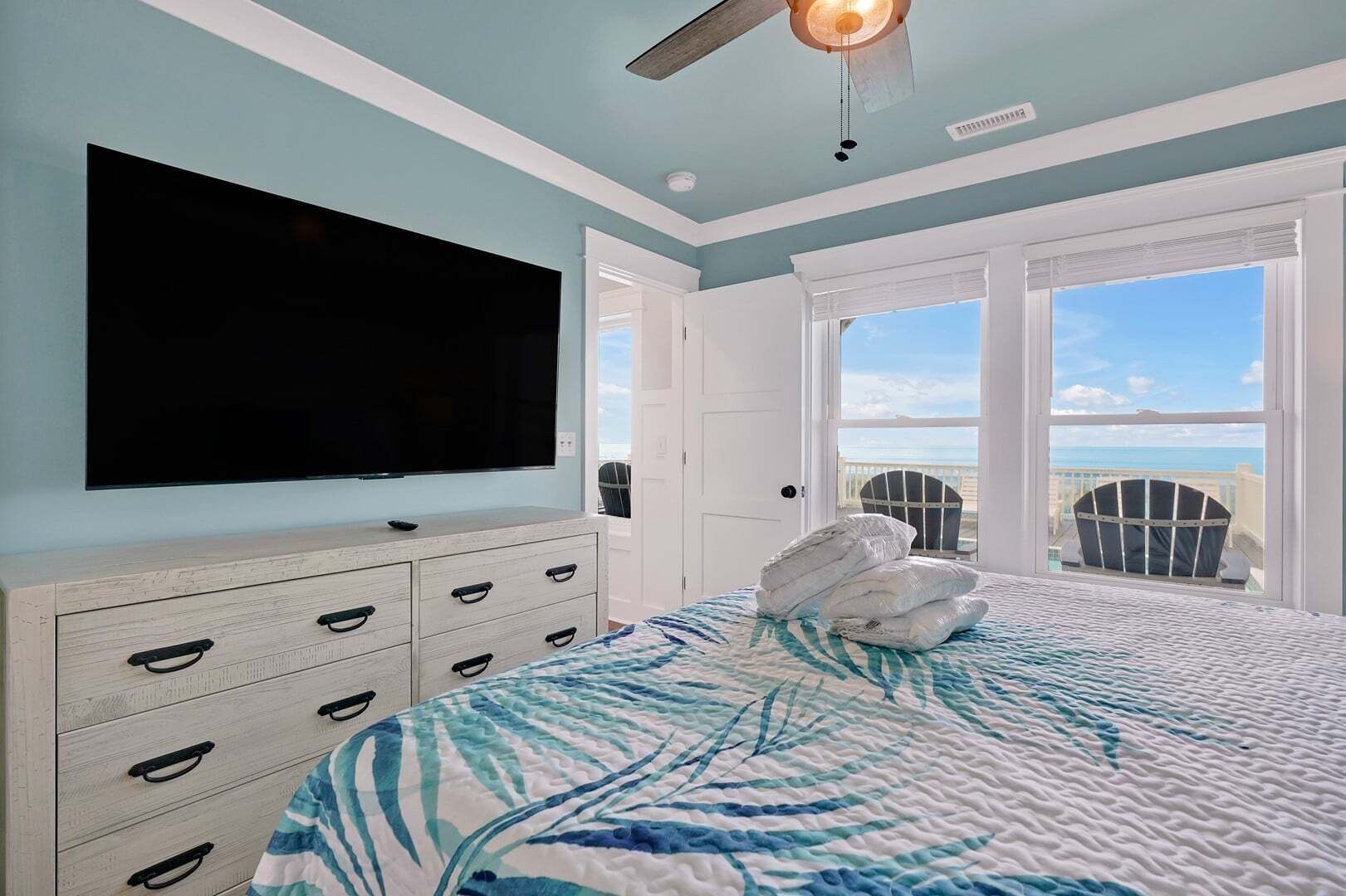 A bedroom with a large TV, a bed with a blue and white patterned bedspread, a wooden dresser with drawers, and a view of the ocean through the window.
