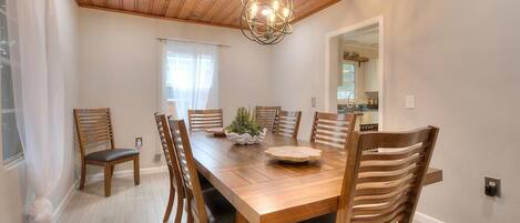 A dining room with a wooden table and chairs, a chandelier, and a window with curtains.