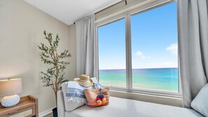 Ein Zimmer mit einem großen Fenster und Blick auf den Ozean, eine Couch mit Hut und bunter Tasche, eine Lampe und eine kleine Pflanze.