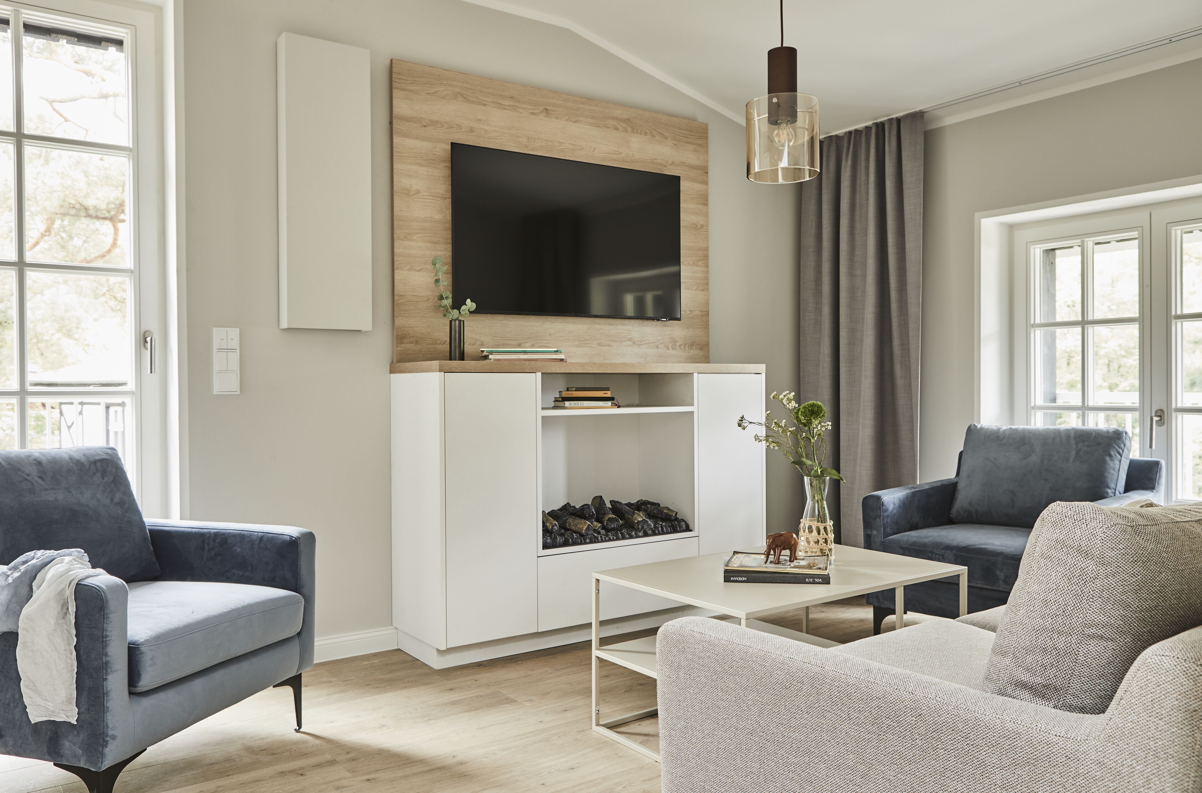A modern living room with a flat-screen TV mounted on a wooden panel, a white entertainment unit with books and decorative items, two sofas, a coffee table, and a large window with curtains.