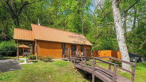 A wooden cabin with a metal roof, a covered porch, and a wooden bridge over a stream. - Studio log cabin-convenient -pet friendly-hot tub- near PF parkway! (Pigeon Forge)
