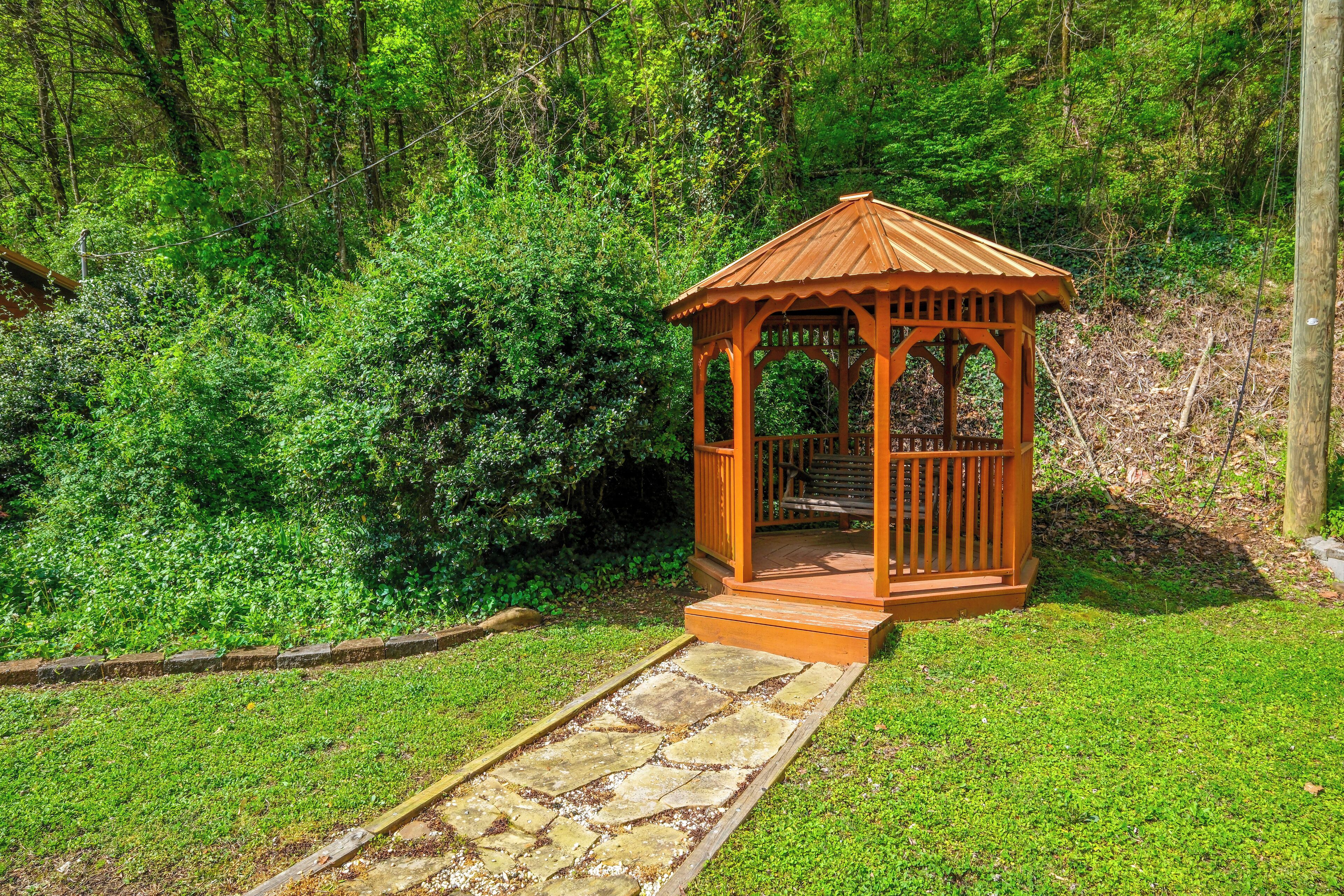 A wooden gazebo with a stone pathway leading to it, surrounded by lush greenery.