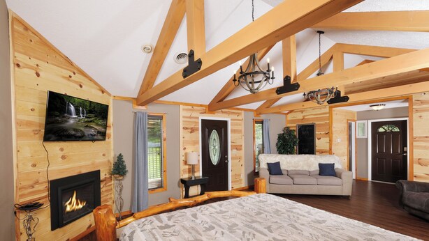 A spacious room with a large bed, wooden beams, a fireplace, a television, and a cozy seating area. - Studio log cabin-convenient -pet friendly-hot tub- near PF parkway! (Pigeon Forge)