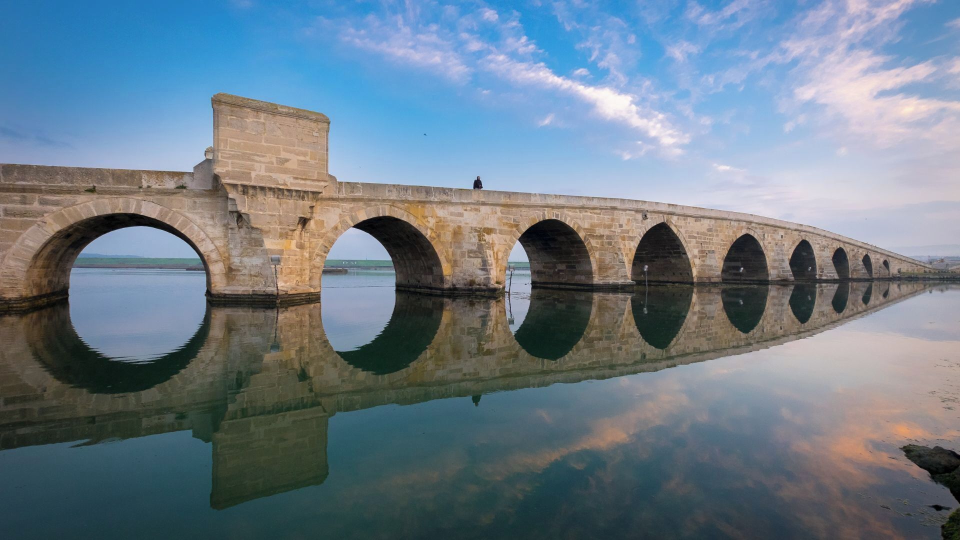 A stone bridge with multiple arches over a calm body of water, reflecting the structure and sky.