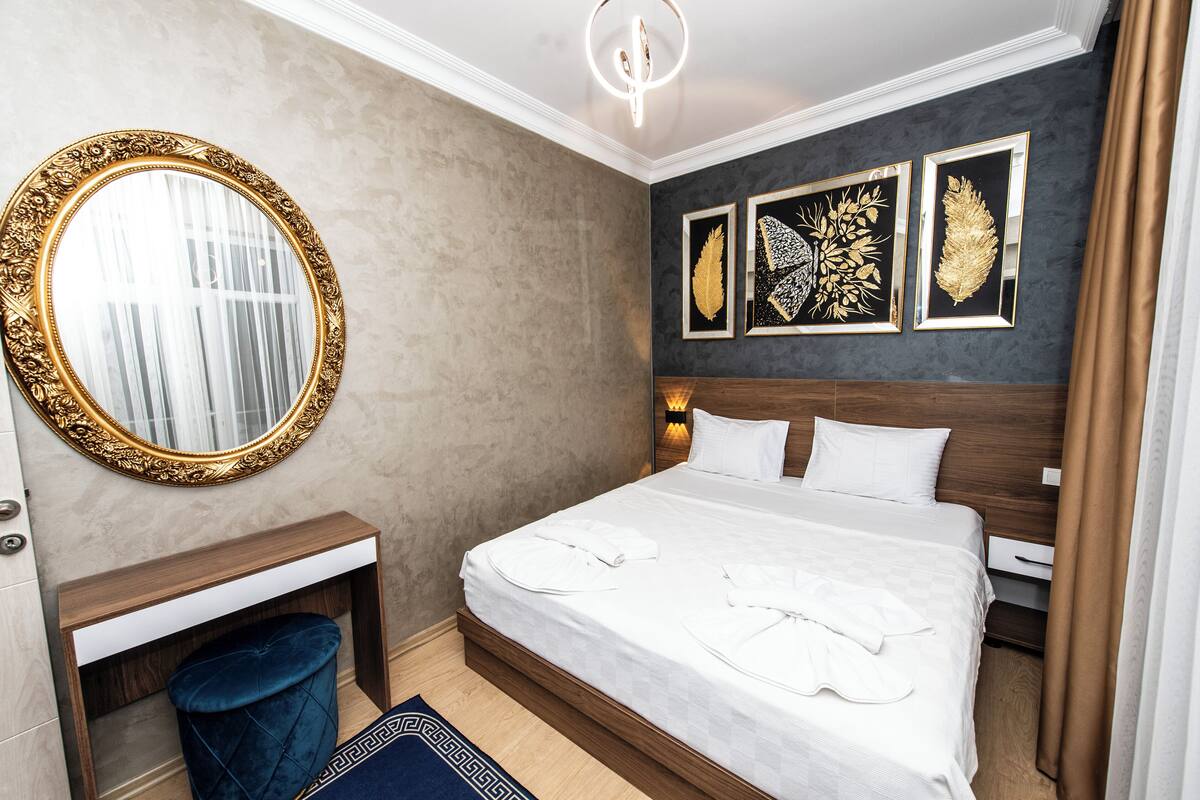 Siististi järjestetty hotellihuone, jossa on suuri soikea peili, valkoisilla lakanoiden peitetty sänky, yöpöytä ja pieni sininen rahi.
