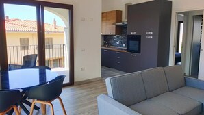 A modern apartment with a dining area, a gray sofa, and a kitchen with dark cabinets and a built-in oven. - Suite Apartments i4MORI Porto Pino (Sant'Anna Arresi)