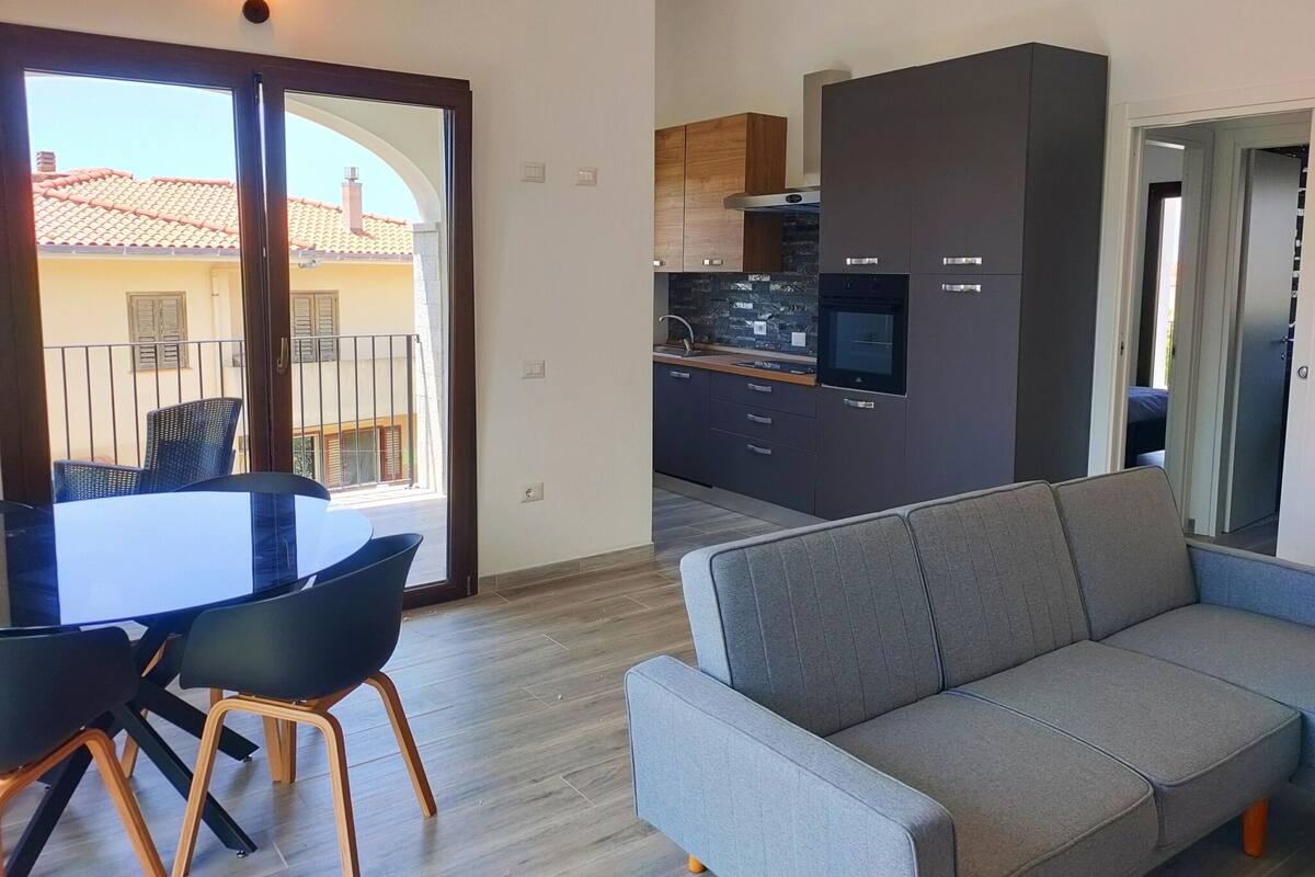 A modern apartment with a dining area, a gray sofa, and a kitchen with dark cabinets and a built-in oven.