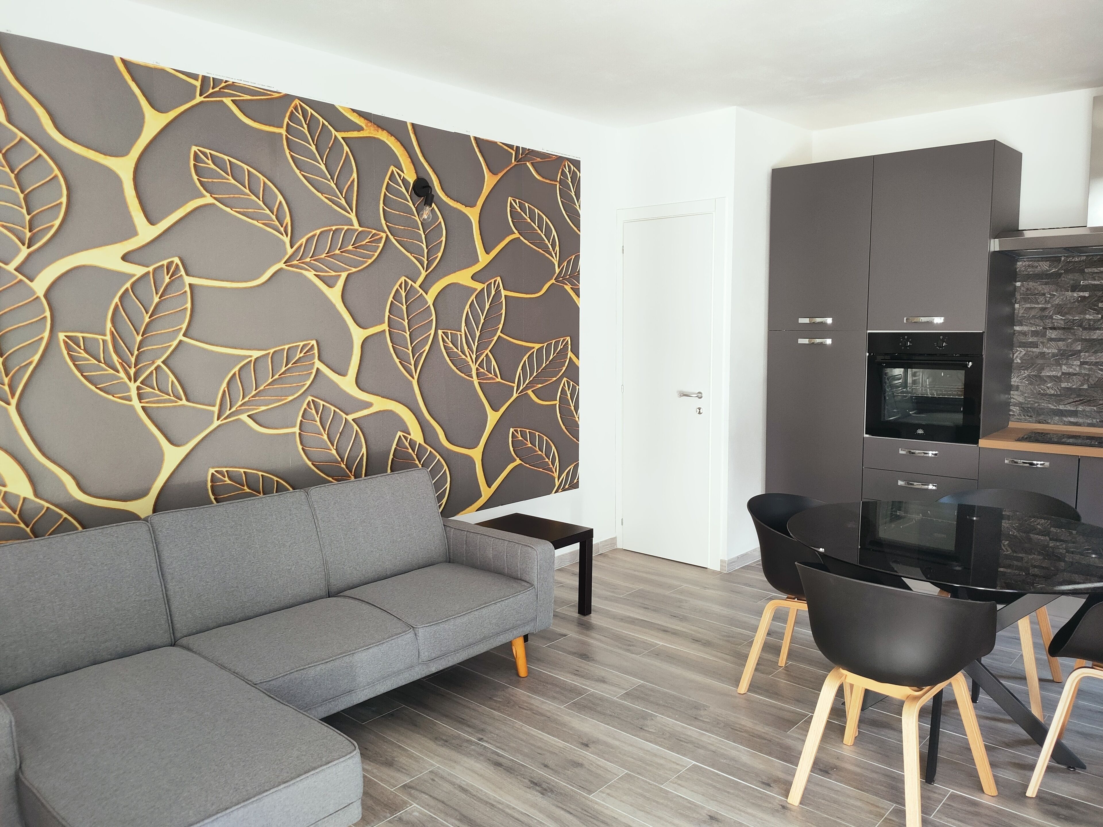 Uma sala de estar moderna com sofá cinza, mesa de jantar preta e cadeiras, área de cozinha com armários cinzas e forno preto, e uma parede decorativa com padrão de folha.