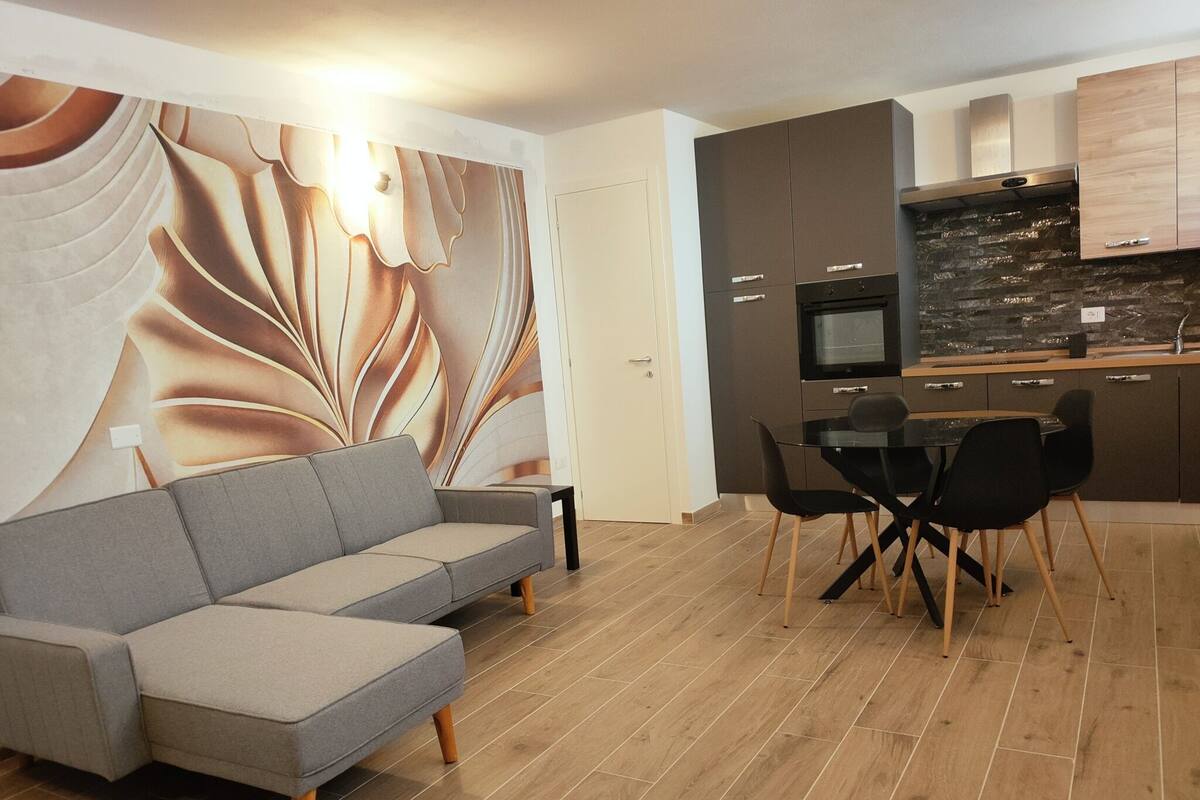 A modern living room with a grey sofa, a dining table with black chairs, and a kitchen area with dark cabinets and a stone backsplash.