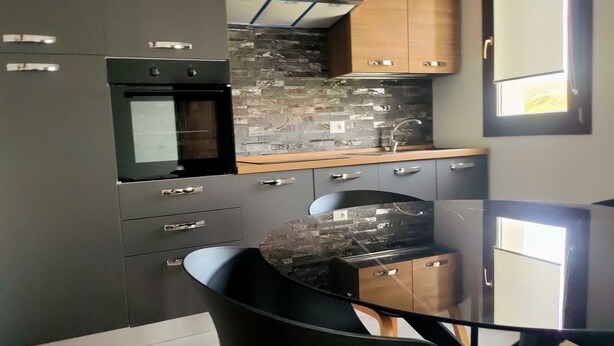 A modern kitchen with dark cabinetry, a built-in oven, a stone backsplash, and a window. - Suite Apartments i4MORI Porto Pino (Sant'Anna Arresi)