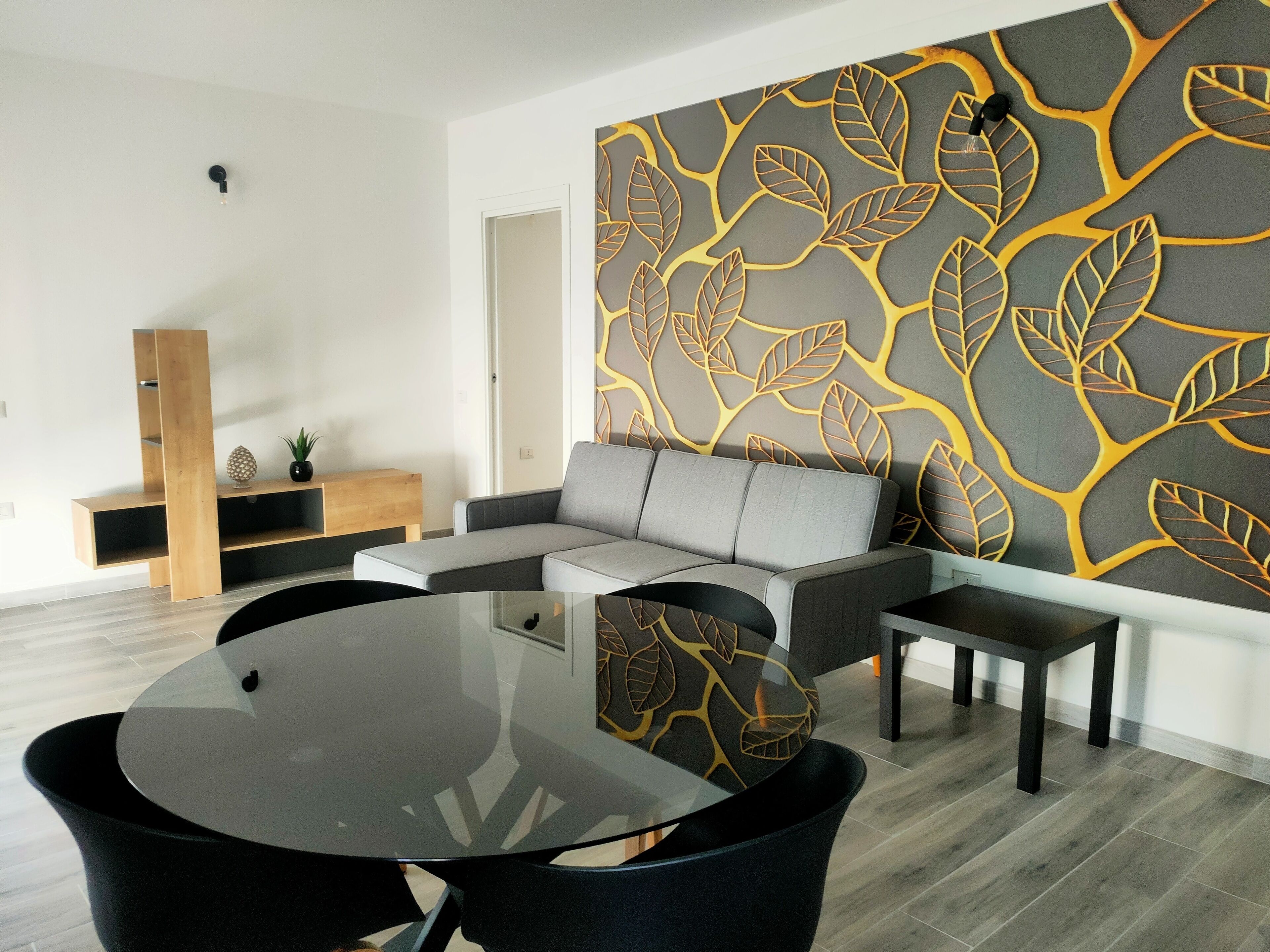 A modern living room with a grey sectional sofa, a glass coffee table, and black chairs. There is a wooden entertainment unit with a plant and decorative items. The wall features a leaf-patterned design.