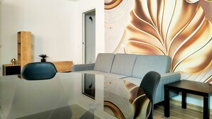 A modern living room with a glass coffee table, grey sofa, and a black armchair. A wooden cabinet and a small plant are visible in the corner. - Suite Apartments i4MORI Porto Pino (Sant'Anna Arresi)