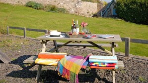 A picnic setup on a wooden table with a blanket and cushions, a bottle of wine, and a glass of water.