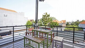 Un balcó amb baranes metàl·liques, taules i bancs de fusta, i vista a edificis i arbres.