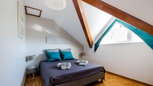Un dormitorio con techo inclinado, una cama con cubrecama oscuro, dos almohadas, una mesita de noche con una lámpara y una ventana con cortina azul turquesa.