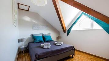 Une chambre avec un plafond en pente, un lit recouvert d’une couverture sombre, deux oreillers, une table de chevet avec une lampe et une fenêtre ornée d’un rideau turquoise.
