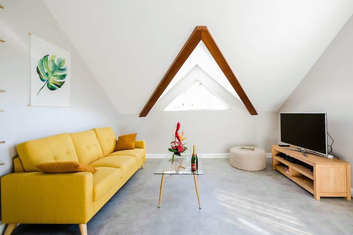 Un salon moderne avec un canapé jaune, une table basse en verre, un meuble TV en bois et une lucarne.