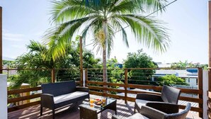 Una terraza de madera con mobiliario de mimbre, una palmera y vistas de edificios al fondo.
