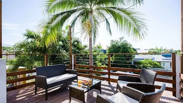 Une terrasse en bois avec des meubles en osier, un palmier et une vue sur des bâtiments en arrière-plan.
