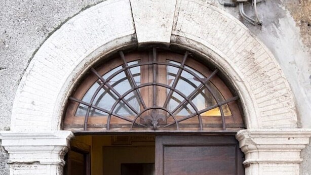 Une porte en bois ornée d’une grille métallique et d’une plaque nominative, encadrée par un arc surmonté de colonnes en pierre.