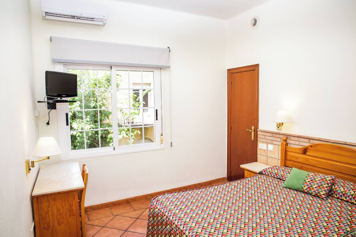 A bedroom with a bed, a desk, a chair, a television, and a window with a view of greenery.