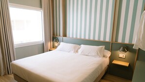 A neatly made bed in a room with striped wallpaper, a bedside table with a lamp, and a window with curtains. - Hotel Can Solé (Cambrils)