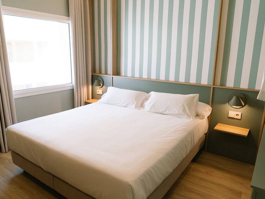 Een keurig opgemaakt bed in een kamer met gestreept behang, een nachtkastje met een lamp en een raam met gordijnen.