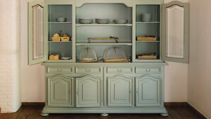 A light green, two-tiered china cabinet with open doors, displaying plates, bowls, and baskets. - Hotel Can Solé (Cambrils)