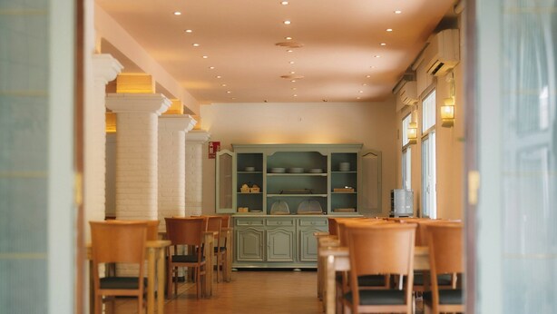 A spacious hallway with wooden chairs and tables, leading to a kitchen area with cabinets and a water dispenser. - Hotel Can Solé (Cambrils)