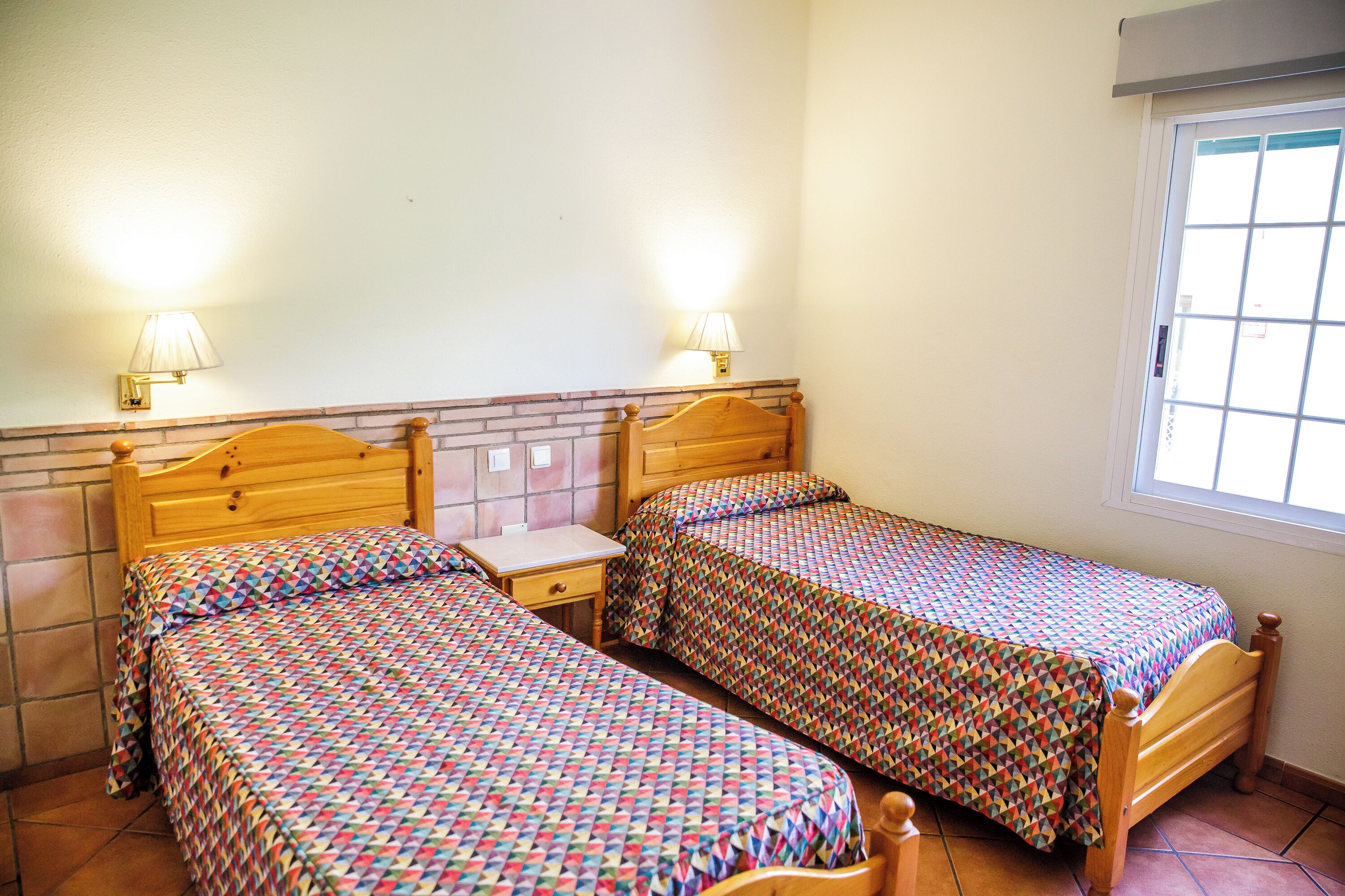 Habitació amb dos llits de fusta, cadascun amb capçal i peu a joc. Hi ha dues taules de nit amb làmpades, un petit calaix i una finestra amb persianes.